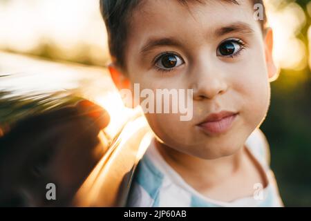 Nahaufnahme eines kleinen lächelnden Jungen, der seinen Kopf aus dem Autofenster streut und die Kamera anschaut. Roadtrip. Kinderleben. Stockfoto