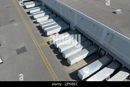 Eine Luftaufnahme über weißen Anhängern, die in einem großen Lagerhaus abgestellt wurden und an den Ladedocks laden. An einem sonnigen Tag aus einer hohen, schrägen Sicht gesehen. Stockfoto