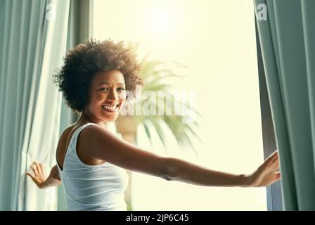 Glückliches Aufwachen zu diesem schönen Tag. Eine attraktive junge Frau öffnet die Vorhänge in ihrem Schlafzimmer zu Hause. Stockfoto