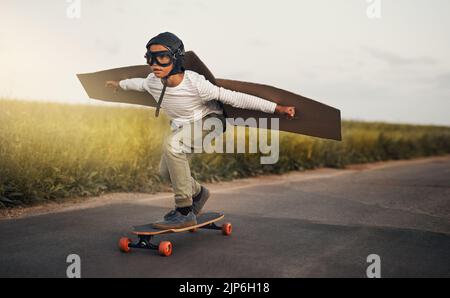 Ein kleiner Junge, der vorgibt, mit einem Paar Pappflügeln zu fliegen, während er draußen auf einem Skateboard reitet. Stockfoto