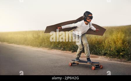 Hes hat die wildeste Phantasie. Ein kleiner Junge, der vorgibt, mit einem Paar Pappflügeln zu fliegen, während er draußen auf einem Skateboard reitet. Stockfoto