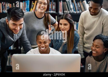 Umgeben Sie sich mit akademisch gesinnten Menschen. Eine Gruppe junger Studenten, die einen Computer zusammen in einer Universitätsbibliothek benutzen. Stockfoto