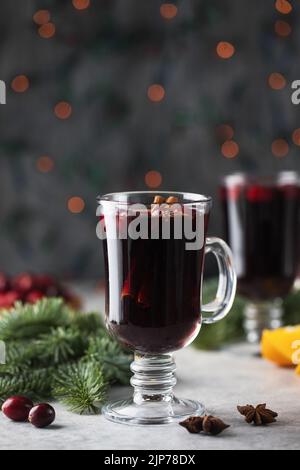 Ein Glas scharf würzigen Glühwein, umgeben von Gewürzen und Zweigen eines Weihnachtsbaums mit Bokeh im Hintergrund. Stockfoto