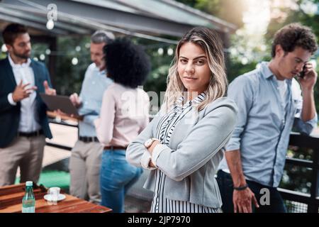 Vertrauen in die Arbeit, die wir tun. Porträt einer selbstbewussten jungen Geschäftsfrau, die draußen mit ihren Kollegen im Hintergrund steht. Stockfoto