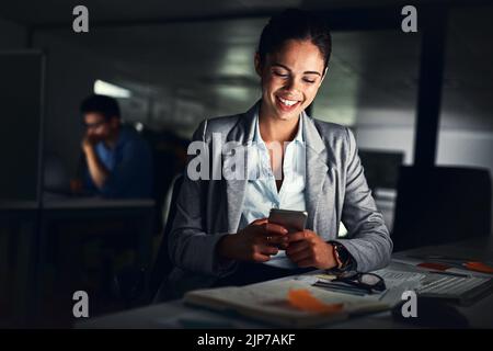 Frau liest spät am Telefon Text, lächelt auf eine Nachricht und schaut sich Online-Beiträge in sozialen Medien an, während sie nachts in einem dunklen Büro arbeitet. Glücklich Stockfoto