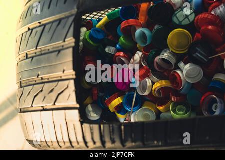 Öffentlicher Ort sammeln in Autoreifen voller bunten Plastikflaschen Becher, eine Handvoll Abfall von Plastikdeckeln. Recycling. Flaschenverschlüsse sammeln. Umweltkonzept Plastikflaschen-Verschluss, der aus gesammelten Flaschen zurückgelassen wird, um in andere Gegenstände zur Wiederverwendung recycelt zu werden Stockfoto