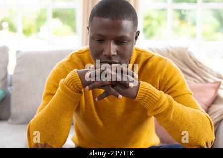 Nachdenklicher afroamerikanischer Mann, der zu Hause auf der Couch sitzt und nach unten schaut und sich auf verschlammten Händen das Kinn ausruht Stockfoto