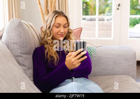 Lächelnde kaukasische Frau, die mit dem Smartphone auf der Couch sitzt Stockfoto