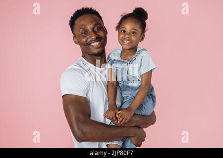 Fröhliches kleines afroamerikanisches Mädchen mit buschigen Haaren, lächelnd auf den Händen von Vater, junger Mann, rosafarbener Studiohintergrund, Panorama mit Kopierraum, Webbanner für Familie, Vaterschaft-Konzept. Stockfoto