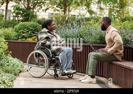 Seitenansicht Porträt einer schwarzen Frau im Rollstuhl im Gespräch mit einem Freund oder Partner im Stadtgarten Stockfoto
