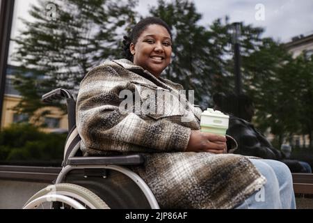 Porträt einer jungen schwarzen Frau im Rollstuhl, die in der Stadt mit der Kamera lächelt und eine Tasse Kaffee hält Stockfoto