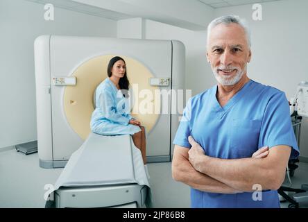Porträt eines erfahrenen Arztes, der mit CT-Scanner im medizinischen Radiologieraum steht, einer Patientin, die hinter seinem Patienten sitzt und auf einen CT-Scan wartet Stockfoto