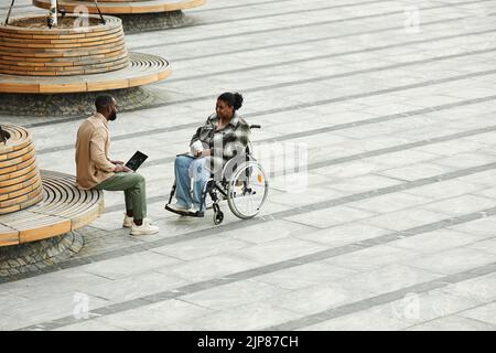 Grafische Weitwinkelansicht eines erwachsenen Paares mit schwarzer Frau im Rollstuhl, das im Freien in der Stadt vor gefliestem Boden plaudert und Platz zum Kopieren bietet Stockfoto