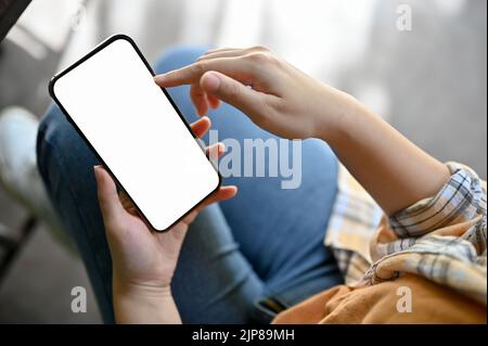 Über dem Kopf geschossen, Eine weibliche Hand hält ein Smartphone weißen Bildschirm Mockup über ihren Schoß. Menschen- und Technologiekonzept. Stockfoto