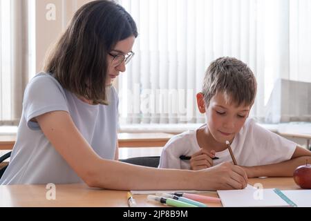 Ein Junge und eine Lehrerin sitzen an einem Tisch mit Heften und Stiften. Die Mutter beobachtet, wie ihr Sohn in einem Notizbuch schreibt. Der Babysitter hilft, s abzuschließen Stockfoto