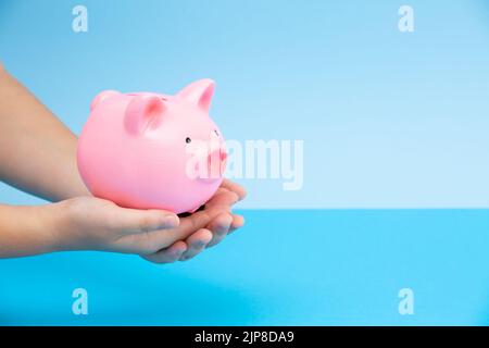 Die Hände der Kinder halten ein rosa Sparschwein in den Händen auf blauem Hintergrund.das Konzept der Investition in die Zukunft des Kindes, Ersparnisse, finanzielle Stockfoto