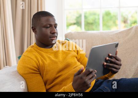 Glücklicher afroamerikanischer Mann, der zu Hause auf der Couch liegt und Tablet sieht Stockfoto