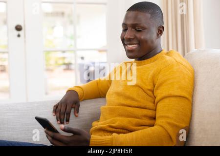 Lächelnder afroamerikanischer Mann, der zu Hause mit dem Smartphone auf der Couch sitzt Stockfoto