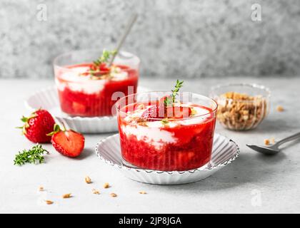 Zwei Gläser mit kaltem Sommerdessert. Erdbeersauce und Joghurtcreme mit frischen Beeren, Müsli und Zitronen-Thymianblättern garnieren. Homemad Stockfoto