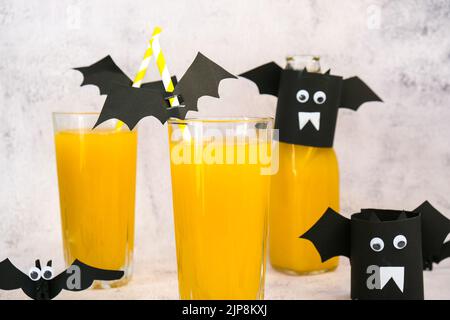 Halloween-Mocktails. Oranger alkoholfreier Cocktail für Halloween-Party auf weißem Tisch. Die Trinkhalme sind mit geschnittenen Papierfledermäusen verziert. Stockfoto