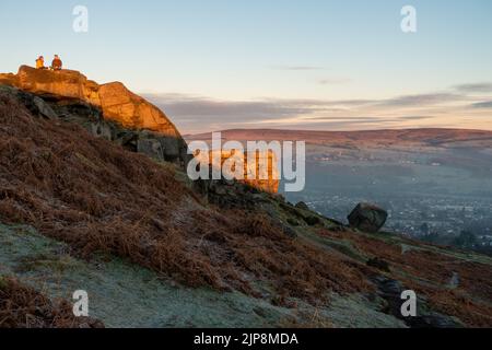 Ein Paar genießt ein heißes Getränk in der goldenen Stunde des Sonnenaufgangs bei The Cow and Calf Rocks, Ilkley Moor, England. Stockfoto