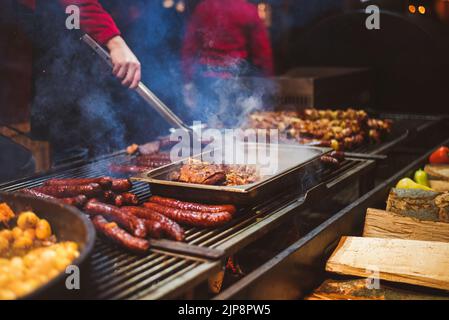 Straßenessen im Freien. Chef Kochen Würstchen, Fleisch und Kartoffeln Kochen auf einem Grill im Freien Stockfoto