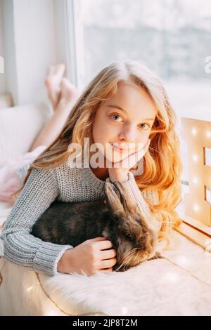 Ein schönes blondes Mädchen sitzt auf dem Fensterbrett mit einem grauen Kaninchen in weihnachtlicher Atmosphäre. Das Konzept der Tierpflege. Stockfoto