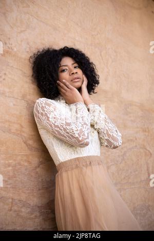 Von unten anmutige junge afroamerikanische weibliche Modell mit dunklen lockigen Haaren in romantischen Outfit berühren Wangen und Blick auf die Kamera, während gelehnt Stockfoto