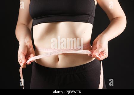 Eine Frau misst den Umfang ihres Bauches auf schwarzem Hintergrund. Das Konzept der Gewichtsabnahme Stockfoto