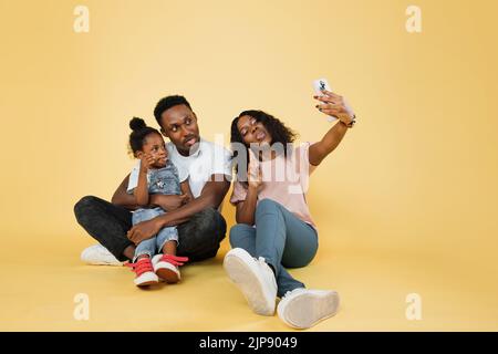 Porträt einer lächelnden afroamerikanischen dreiköpfigen Familie, die mit dem Smartphone auf dem Boden sitzt, während sie ein lustiges Bild von ihnen macht, das Zungen auf gelbem Studiohintergrund zeigt, freien Kopierraum, Banner. Stockfoto