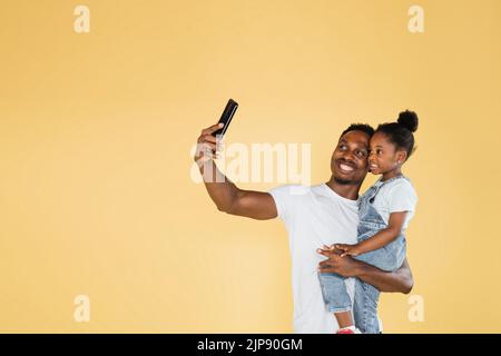 Glückliche afroamerikanische Familie Vater und Tochter Spaß haben und mit Handy-Videoanruf auf gelbem Hintergrund. Selbst, isoliert, soziale Distanzierung, Quarantäne zur Coronavirus-Prävention. Stockfoto