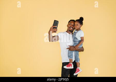 Glückliche afroamerikanische Familie Vater und Tochter Spaß haben und mit Handy-Videoanruf auf gelbem Hintergrund. Selbst, isoliert, soziale Distanzierung, Quarantäne zur Coronavirus-Prävention. Stockfoto