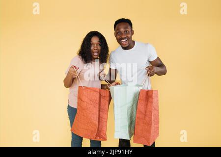 Aufgeregte afroamerikanische Frau und Mann schauen sich die Kamera an und halten Einkaufstaschen. Glücklich lockig Mädchen und Kerl in lässigen Kleidung glücklich, dass die Käufe. Stockfoto