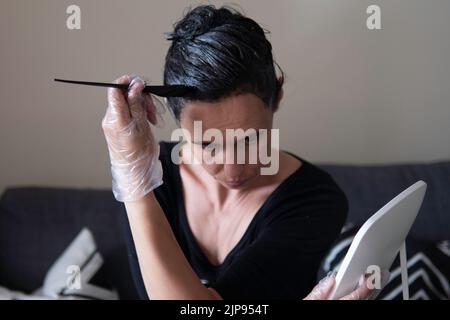 Frau mittleren Alters Färbung dunkle Haare mit grauen Wurzeln zu Hause. Frau, die auf der Couch sitzt und mit einem schwarzen Pinsel Haare färben und zum Spiegel schaut Stockfoto