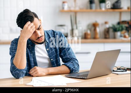 Trauriger arabischer oder indischer Mann, der an einem Schreibtisch sitzt, müde und gestresst aussieht, frustriert auf Arbeitsgrafiken schaut, die Arbeit am Projekt nicht rechtzeitig beendet, braucht Ruhe. Arbeitsüberlastung, Termin Stockfoto