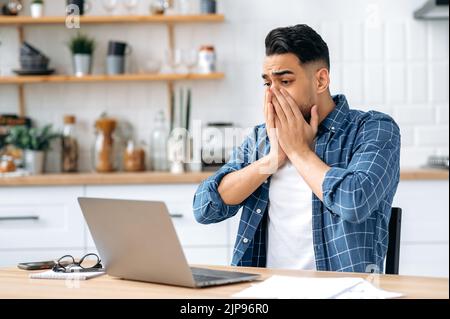 Schockiert besorgter arabischer oder indischer Mann, sitzt am Schreibtisch in der Küche, benutzt Laptop für Online-Arbeiten, schaut überrascht auf den Bildschirm, bedeckt seinen Mund mit den Händen, scheiterte den Deal, erlebt Stress Stockfoto