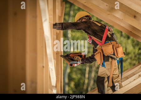 Kaukasischer Arbeiter mit Sicherheitsgeschirr und Schutzhelm auf der Baustelle des hölzernen Wohngebäudes. Arbeiten in der Dachsektion, Drivin Stockfoto