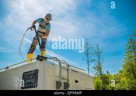 Kaukasischer Mann in Uniform und gelben Gummistiefeln auf dem Dach des weißen RV stehend Reinigung der Oberfläche mit einer großen Auto-Waschbürste. Sauberes Blue Sky und Stockfoto