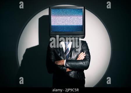 Mann mit Fernseher statt Kopf. Media Zombie-Konzept mit fernsehsüchtiger Rüde im schwarzen Mantel mit gekreuzten Armen, die im Rampenlicht im Studio posieren. Kontra Stockfoto