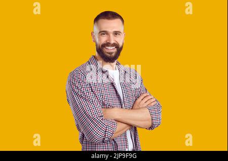 Fröhlicher junger Mann in einem lässigen Hemd, der mit gefalteten Armen auf einem gelben Studiohintergrund steht Stockfoto