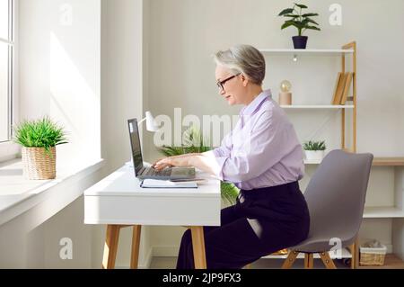 Reife Geschäftsfrau, die an ihrem Schreibtisch in einem modernen Bürogebäude an ihrem Laptop arbeitet Stockfoto