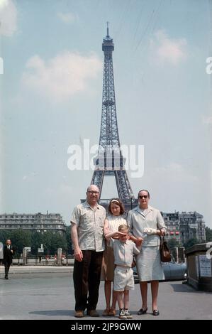 1964, eine historische vierköpfige Familie, die Paris, Frankreich, besucht und für ihr Foto mit dem ikonischen Wahrzeichen, dem Eiffelturm, auf dem Bild hinter ihnen steht. Erbaut, um eine der Hauptattraktionen auf der Weltausstellung der Stadt im Jahr 1889 zu sein, lag sein Fokus auf den riesigen Konstruktionen aus Eisen und Stahl, die den großen industriellen Fortschritt dieser Zeit waren. Wie viele Menschen haben vor dem berühmten französischen Wahrzeichen gestanden, um Fotos zu machen? Stockfoto