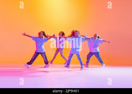 Gruppe von Kindern, kleine Mädchen in sportlichen Casual-Stil Kleidung tanzen in Choreographie Klasse isoliert auf orangefarbenem Hintergrund in lila Neonlicht. Stockfoto