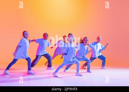 Hip-Hop-Tanz, Street-Style. Glückliche Kinder, kleine aktive Mädchen in lässiger Kleidung tanzen isoliert auf orangefarbenem Hintergrund in purpurem Neonlicht. Stockfoto