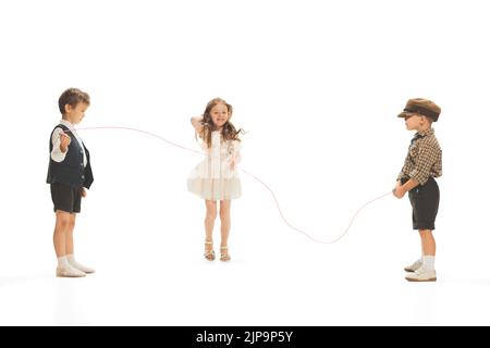 Portrait von drei Kindern, Jungen und Mädchen in Retro-Kleidung, die zusammen spielen und über ein isoliertes Seil über weißen Studiohintergrund springen Stockfoto