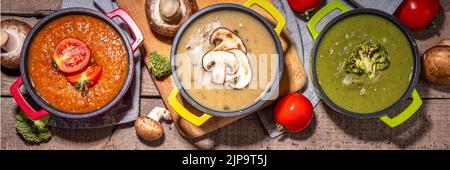 Herbstgemüsecremesuppen Set. Drei Portionstöpfe mit verschiedenen veganen heißen Gemüsesuppen – Tomaten, Pilze, Brokkoli auf rustikalem Holztisch Stockfoto