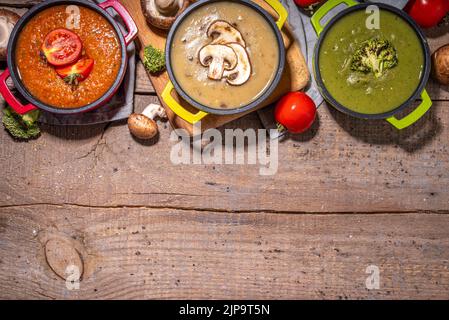 Herbstgemüsecremesuppen Set. Drei Portionstöpfe mit verschiedenen veganen heißen Gemüsesuppen – Tomaten, Pilze, Brokkoli auf rustikalem Holztisch Stockfoto