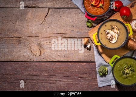 Herbstgemüsecremesuppen Set. Drei Portionstöpfe mit verschiedenen veganen heißen Gemüsesuppen – Tomaten, Pilze, Brokkoli auf rustikalem Holztisch Stockfoto