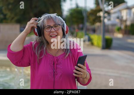 Ältere Frau mit weißen Haaren und Brille, mit ihrem Handy und Kopfhörern, die versuchen, jedem zuzuhören, der mit ihr spricht. Stockfoto