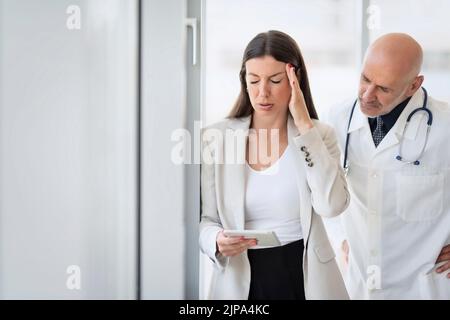 Geschäftsfrau, die am Fenster im Büro steht und Migräne hat. Männlicher Arzt, der neben ihr steht. Stockfoto